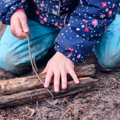Closeup of low section of a caucasian child girl in warm winter clothing kneeing on the forest ground and playing with a piece of wood and a string and trying to make a knot. Seen in Germany in January
